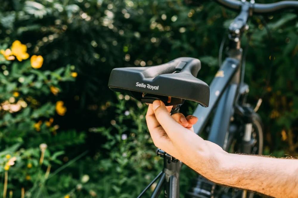 Schwarzer Selle Royal Sattel Vaia Moderate Unisex, von Hand gehalten, vor verschwommenem Fahrrad und grüner Natur.
