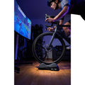 ELITE Sterzo Smart Travelblock Vorderradstütze mit Lenkfunktion für Radfahrer beim Indoor-Training vor großem Bildschirm.
