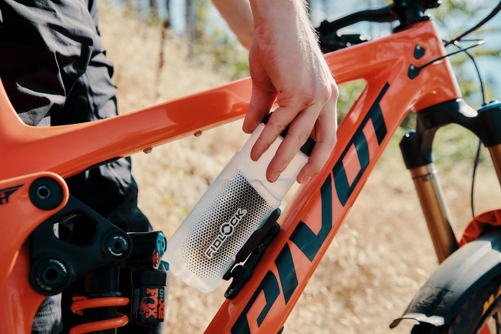 FIDLOCK TWIST Trinkflasche an magnetischer Halterung am orangefarbenen Mountainbike wird von Hand bedient.