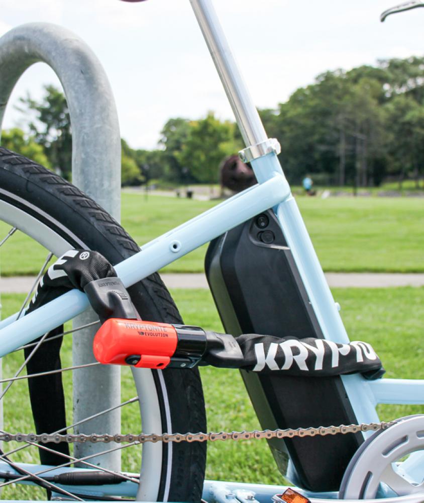 Kryptonite Evolution 4 I.C. Kettenschloss sichert hellblaues E-Bike an Fahrradständer. Schwarz mit orangefarbenem Schloss.
