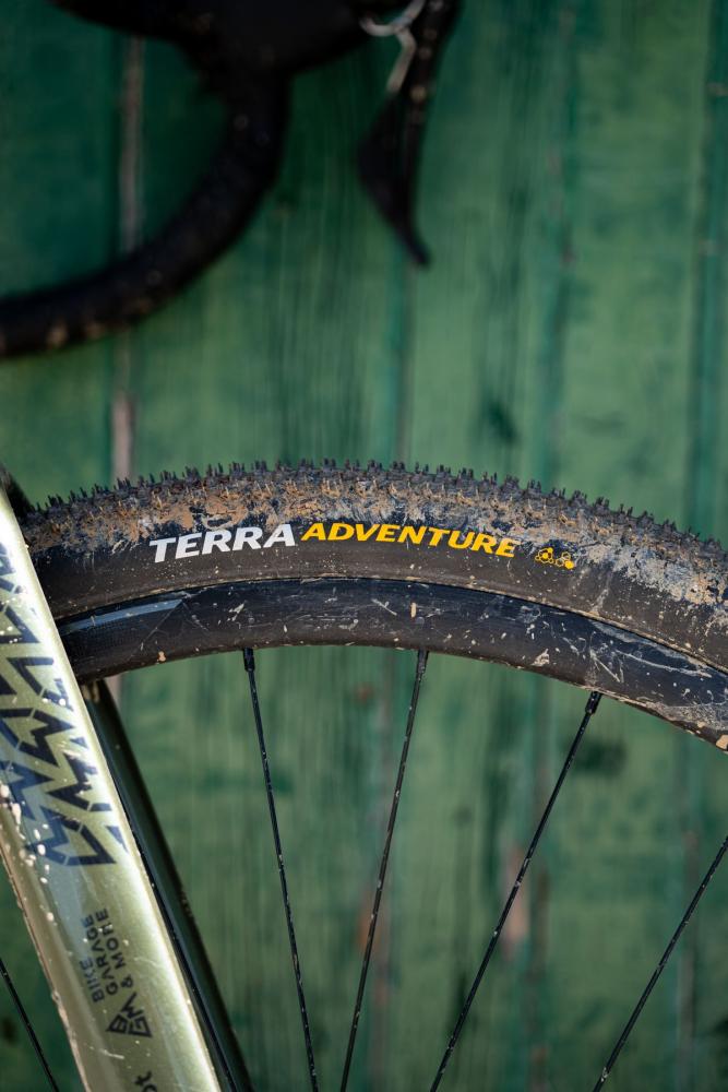 Continental Terra Adventure Gravel & Trail Reifen auf schmutzigem Fahrradrad vor grüner Holzwand.