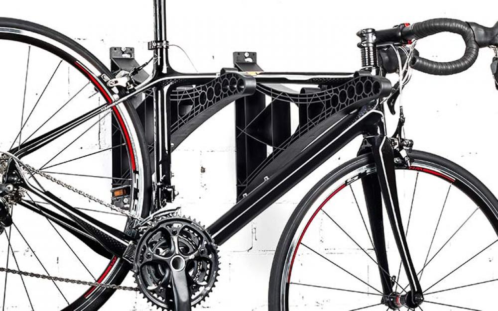 Schwarzes Rennrad an einer modernen, schwarzen Wandhalterung mit Gittermuster vor weißer Ziegelwand.
